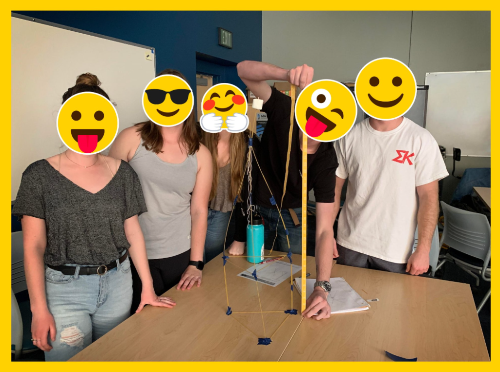 Five people with emoji faces stand around a table in a classroom. On the table in front of them is a tall, makeshift structure made of yellow straws and blue tape. One person is holding the top of the structure. Everyone appears to be enjoying themselves, showcasing their creativity in an entrepreneurship curriculum activity.